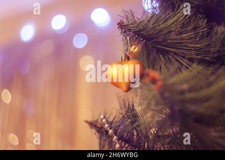 Goldenes Weihnachtsspielzeug Herz auf einem Fichtenzweig auf einem glänzenden Hintergrund mit Bokeh, selektiver Fokus Unschärfe Hintergrund Kopie Raum. Stockfoto