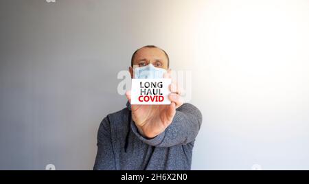 COVID-19 Symbol für langstreckige kovide Symptome. Weiße Karte, Worte Langstrecke covid. Ein junger Mann in grauer Kleidung und medizinischer Maske. Schöner weißer Hintergrund. Stockfoto