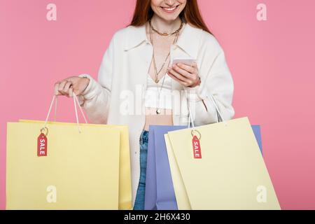 Verkürzte Ansicht der stilvollen Frau mit Einkaufstaschen mit Smartphone isoliert auf rosa Stockfoto