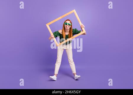 In voller Größe Foto von lustigen roten Haaren junge Dame mit Rahmen tragen Brille Bluse Hose isoliert auf violettem Hintergrund Stockfoto