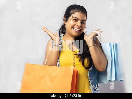 Eine schöne indische Frau in gelbem Kleid mit Einkaufstaschen, die auf weißem Hintergrund auf die Kamera schaut Stockfoto