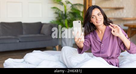 Foto einer Frau mittleren Alters liegt im Bett mit Handy. Sie wacht auf und macht Selfie für Social-Media-Beiträge oder Blogs. Moderne Technologie und digitale süchtig Stockfoto