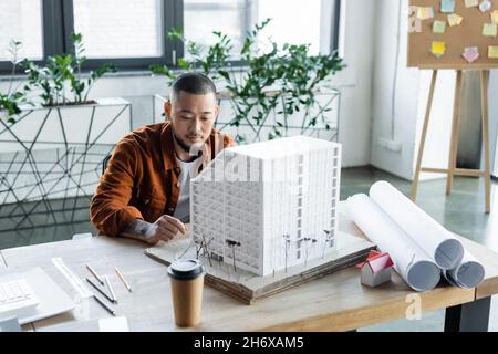 asiatischer Architekt, der in der Nähe des Hausmodells sitzt, während er an einem Architekturprojekt im Büro arbeitet Stockfoto