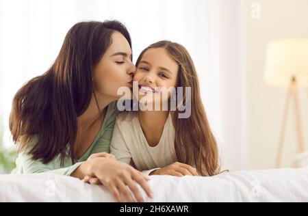 Glückliche Mutter küsst ihre hübsche kleine Tochter, während sie sich zu Hause auf einem Bett kuschelt Stockfoto