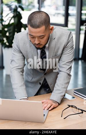 asiatischer Geschäftsmann, der im Büro am Schreibtisch steht und einen Laptop benutzt Stockfoto