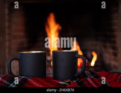 Kaffeetassen auf einem Sofa rot karierte Decke, brennender Kamin Hintergrund. Weihnachtsurlaub gemütlich warmes Zuhause. Zwei schwarze Teebecher, Entspannung am Feuer Stockfoto