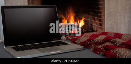 Winter Home Office-Konzept. Leerer Bildschirm Laptop auf brennendem Kamin Hintergrund, schwarzer leerer Computerbildschirm, gemütliches Zimmer innen. Weihnachten warm hous Stockfoto