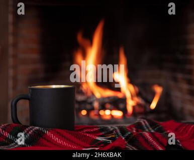 Weihnachtsurlaub gemütlich warmes Zuhause. Tasse Kaffee auf einem Sofa rot karierte Decke, brennender Kamin Hintergrund. Schwarzer Teebecher, Entspannung am Feuer Stockfoto