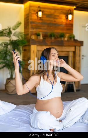 Das vertikale Foto einer Schwangeren sitzt auf einem Bett und genießt ihre Lieblingsmusik. Sie hört die Musik über die Online-Smartphone-Anwendung. Modern Stockfoto