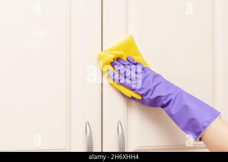 Weibliche Hand in einem violetten Gummihandschuh wischt die beigefarbene Tür des Küchenschranks ab. Gelber Lappen in der Hand Stockfoto