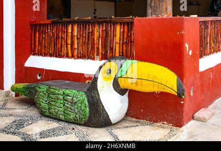 Holzbank in Tukan-Form geschnitzt sitzt auf mosiac Bürgersteig gegen bambo und Stuckwand in Mexiko Stockfoto