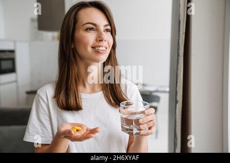 Lächelnde junge Brünette Frau hält Pillen und ein Glas Wasser, während sie zu Hause standing Stockfoto
