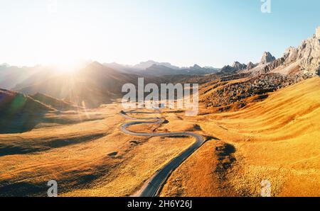 Unglaubliche Luftaufnahme auf kurvenreicher Straße im herbstlichen Bergtal bei Sonnenuntergang. Das goldene Abendlicht beleuchtet die Berge und das orangefarbene Gras. Giau-Pass, Dolomiten, Dolomiten, Italien Stockfoto