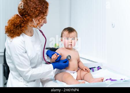 Eine rothaarige Ärztin untersucht das kleine Kind weniger als ein Jahr lang in einer modernen Klinik. Kinderarzt während der Untersuchung Baby im Krankenhaus Stockfoto