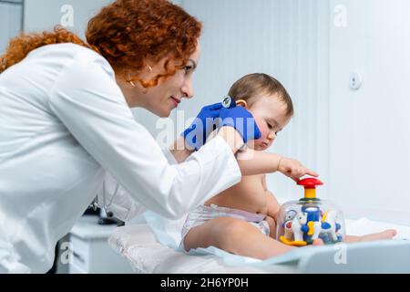 Eine rothaarige Ärztin untersucht das kleine Kind weniger als ein Jahr lang in einer modernen Klinik. Kinderarzt während der Untersuchung Baby im Krankenhaus Stockfoto