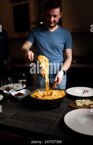 Mann stellt den Tisch, hausgemachte Pasta, Speck, Feta und Tomaten, Käse mit blauem Schimmel, Wein. Tiefe Teller für Pasta. Draufsicht. Vertikales Foto. Stockfoto