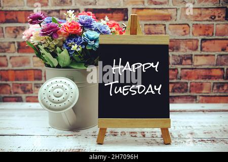 Happy Tuesday Text auf schwarzer HolzStaffelei und Vase mit Blumenstrauß auf Backsteinwand Hintergrund Stockfoto