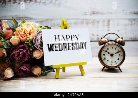 Happy Wednesday Text mit Blumenstrauß und Wecker auf Holzhintergrund Stockfoto