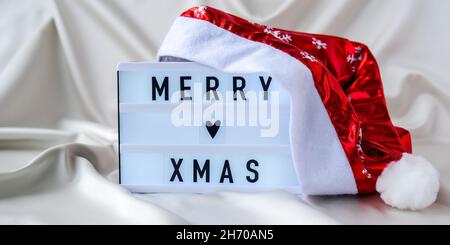 Lightbox mit Text FROHE WEIHNACHTEN mit weihnachtsmütze auf Seidenstoff-Hintergrund. Konzept für Winterurlaub. Stockfoto