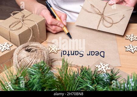 Eine Frau bittet den Weihnachtsmann um Glück. Weibliche Hand schreibt einen Weihnachtsbrief an den Weihnachtsmann, hält einen Umschlag auf dem Tisch mit Weihnachtsgeschenken, Tannenbäumen und Stockfoto