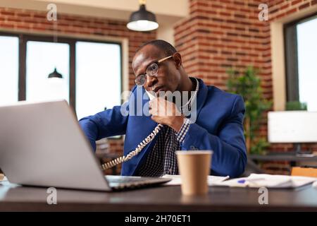 afroamerikanischer Mann, der Bürotelefon benutzte, um über Geschäftsstrategie und Projekt zu sprechen. Ein Mitarbeiter des Unternehmens telefoniert mit dem Manager, um das Projekt zu planen, und arbeitet mit dem Festnetztelefon. Stockfoto