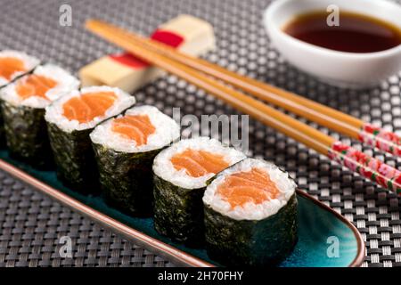 Lachs-Hosomaki-Sushi, serviert im Restaurant, mit Essstäbchen und Sojasauce, aus nächster Nähe auf einer gefroteten Bambusmatte Stockfoto