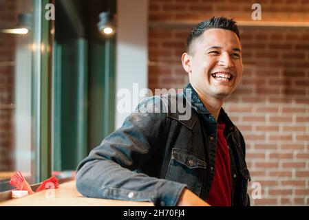Fröhlicher junger hispanischer Kerl in Denim-Jacke, der hell lächelt, während er sich in einem Café im Loft-Stil mit Backsteinmauer in der Nähe des Fensters ausruhte Stockfoto