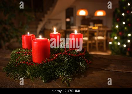 Geschmückter Adventskranz aus Tannenzweigen mit roten brennenden Kerzen auf einem dunklen Holztisch, Wohnzimmer mit im Hintergrund verschwommenem Weihnachtsbaum, Stockfoto