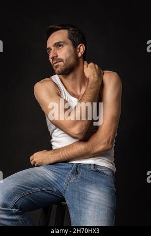 Klassisches Porträt eines jungen hübschen bärtigen traurigen Mannes in weißem Unterhemd und Jeans isoliert auf dunklem Hintergrund. Emotionen, Gesichtsausdruck Stockfoto
