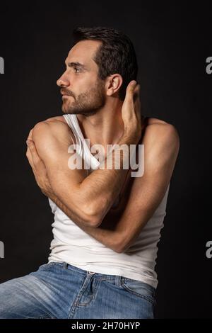Porträt eines jungen, gutaussehenden, bärtigen, traurigen Mannes in weißem Unterhemd und Jeans, der isoliert auf dunklem Hintergrund davonschaut. Emotionen, Gesichtsausdruck Stockfoto