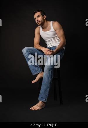 Klassisches Porträt eines jungen hübschen bärtigen traurigen Mannes in weißem Unterhemd und Jeans isoliert auf dunklem Hintergrund. Emotionen, Gesichtsausdruck Stockfoto