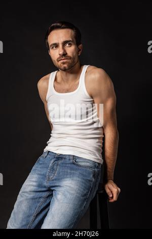Klassisches Porträt eines jungen hübschen bärtigen traurigen Mannes in weißem Unterhemd und Jeans isoliert auf dunklem Hintergrund. Emotionen, Gesichtsausdruck Stockfoto