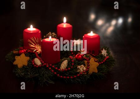 Dekorierter Adventskranz aus Tannenzweigen mit rot angezündeten Kerzen, Weihnachtskugeln und Sternkeksen, einige verschwommene Lichter im dunklen Hintergrund, Copy Spa Stockfoto