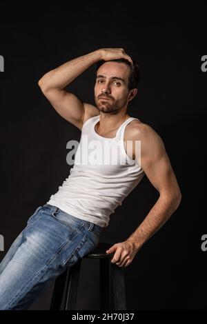 Klassisches Porträt eines jungen hübschen bärtigen traurigen Mannes in weißem Unterhemd und Jeans isoliert auf dunklem Hintergrund. Emotionen, Gesichtsausdruck Stockfoto