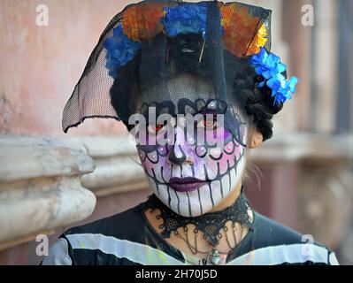 Kostümierte junge Mexikanerin mit gemaltem Gesicht und orangefarbenen Kontaktlinsen trägt am Tag der Toten (Día de los Muertos) einen schwarzen Vogelkäfig-Schleier. Stockfoto