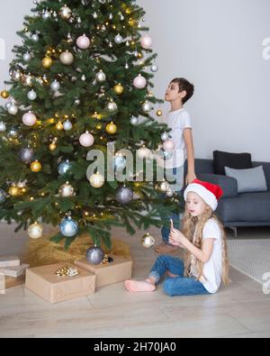 Kinder - ein Junge und ein Mädchen spielen in der Nähe des Weihnachtsbaums. Wohnzimmer mit Weihnachtsbaum und Dekorationen. Neujahr. Geschenk geben. Stockfoto