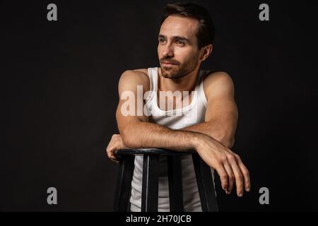 Klassisches Porträt eines jungen hübschen bärtigen traurigen Mannes in weißem Unterhemd und Jeans isoliert auf dunklem Hintergrund. Emotionen, Gesichtsausdruck Stockfoto