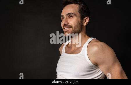 Porträt eines jungen, gutaussehenden, bärtigen, traurigen Mannes in weißem Unterhemd und Jeans, der isoliert auf dunklem Hintergrund posiert. Stockfoto