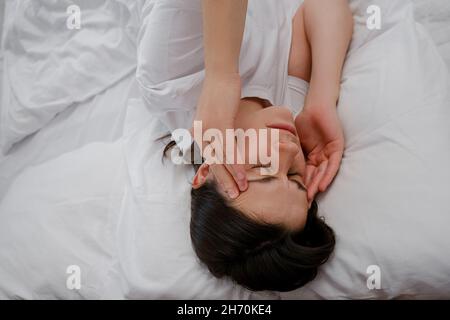 Erschöpfte junge kaukasische Frau, die den Kopf berührt, an starker Kopfschmerzen leidet Migräne, allein auf weißem Bett zu Hause liegend Stockfoto