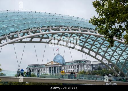 Moderne Glasbrücke des Friedens mit Menschen vor dem Zeremoniellen Palast von Georgien gegen grauen Himmel auf Tiflis