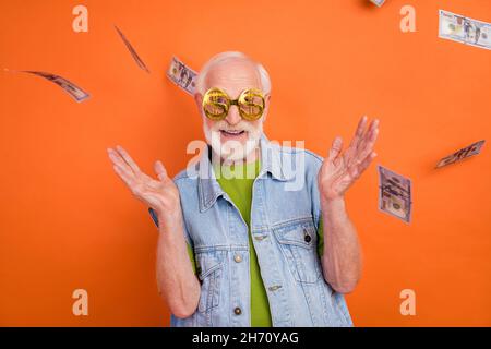 Foto von beeindruckt lustig pensionierten Mann tragen vintage Jeans Weste Geld Gläser Luft Wind weht lächelnd isoliert orange Farbe Hintergrund Stockfoto