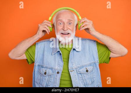 Foto von ziemlich beeindruckt Rentner tragen Vintage Jeans Weste Musik Kopfhörer isoliert orange Farbe Hintergrund hören Stockfoto