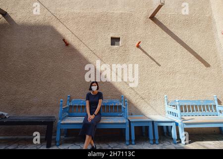 Glückliche Frau in weißer Maske sitzt auf der Bank in der Straße der Bur Dubai Altstadt in der Nähe Creek District. Reisekonzept Stockfoto