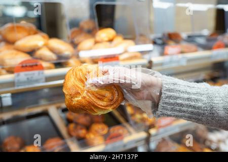 Die Hand einer reifen Käuferin mit Handschuhen hält ein frisches Brötchen, während sie im Supermarkt auf dem Display steht Stockfoto