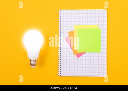 Ein Ideenkonzept - leuchtende Energiesparlampe und EIN Notizblatt mit Haftnotizen auf gelbem Tisch. Draufsicht, Flach Liegend, Leere Vorlage Stockfoto