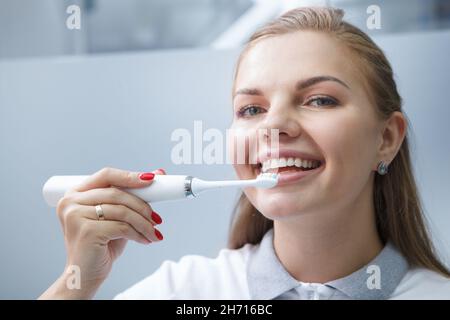 Nahaufnahme einer charmanten jungen Zahnärztin, die lächelt und eine elektrische Zahnbürste an ihren Zähnen verwendet Stockfoto