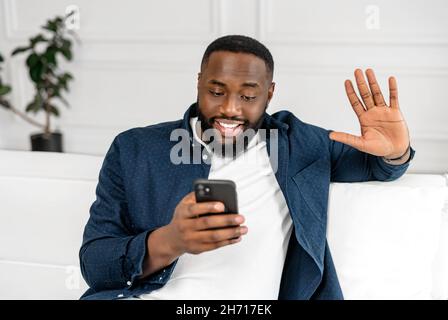 Aufgeregt fröhlich positiv afroamerikanischen Kerl mit toothy Lächeln mit Smartphone für Video-Verbindung, schwarzer Mann in halten Handy und winken, reden auf der Ferne sitzende Sofa zu Hause Stockfoto