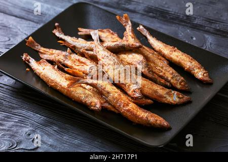 Knusprig gebratener Speer, Shishamo auf einer schwarzen rechteckigen Platte auf einem schwarzen strukturierten Holztisch Stockfoto