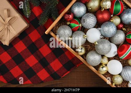 Christmas Flat Lay - Holzkiste mit Weihnachtsbaum-Ornamenten auf einem karierten Trre-Rock mit Geschenk und Kiefernästen. Stockfoto