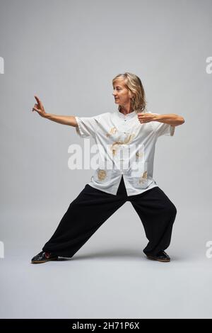 Reife blonde Frau, die Chi Kung und Tai Chi auf weißem Hintergrund praktiziert. Sie trägt eine traditionelle weiße chinesische Tai Chi Jacke, schwarze Hosen und BL Stockfoto